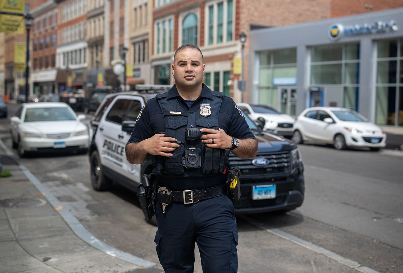 A Waterbury police officer standing in a city street in front of a patrol vehicle