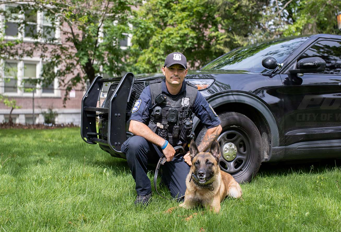 A Waterbury police officer from the K9 unit kneeling next to their police dog in the grass
