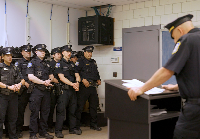 A group of Waterbury police officers stands in a line, listening to another officer who is speaking from behind a podium