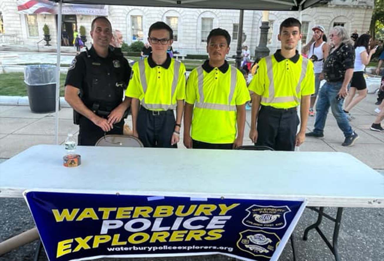 Police officers standing next to three boys in neon shirts behind a table that has a "Waterbury Police Explorers" tapestry