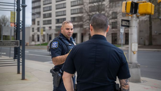 Two Waterbury police officers talking on a city sidewalk near a street intersection